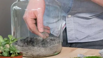 Hand places sand in a large glass container on the table.