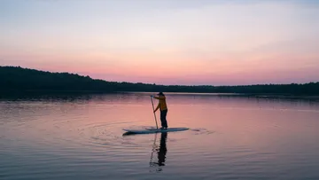 Person paddleboarding on calm lake at sunset.