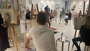 Naked model presents herself as artists sketch. Environment: art class with drawing boards.