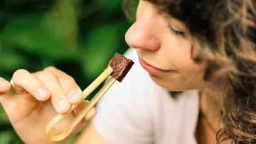 Woman eating chocolate with chopsticks in green surroundings.