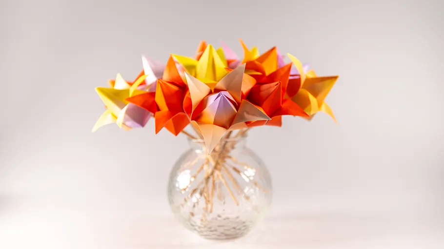 Colourful origami flower arrangement in a glass vase against a neutral background.