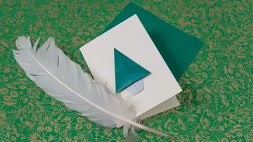 Weißes Blatt mit grünem Baum und Feder auf gemustertem Hintergrund.