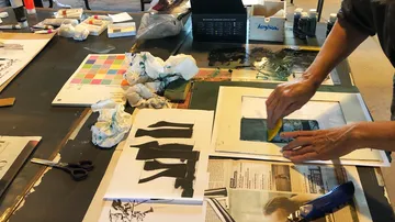 Eine Hand malt mit einer Kelle auf Papier, umgeben von Materialien.