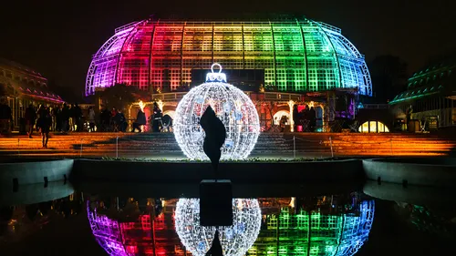 Illuminated greenhouse is reflected in the water, people gather.