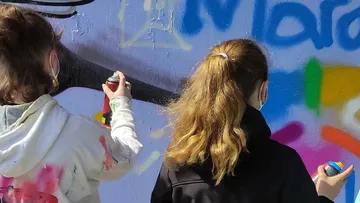 Zwei Personen sprühen Graffiti an eine Wand im Freien.