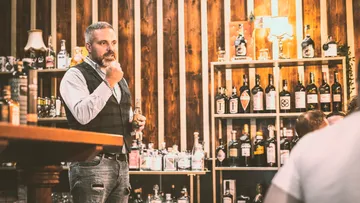Man talking to spirits at bar in front of shelf.