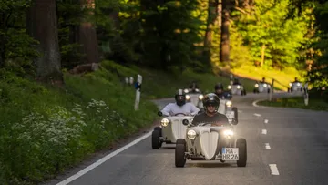 Weißer Sportwagen fährt auf kurvenreicher Landstraße zwischen Bäumen.