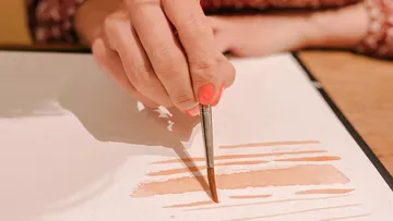 One hand holds a brush over watercolor paper.