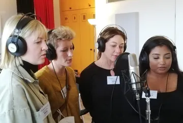 Four women sing in a recording studio with a microphone.