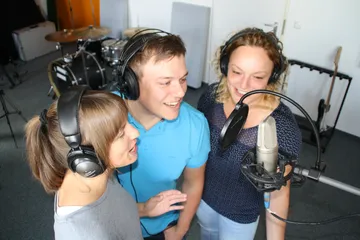 Three people sing together in front of a microphone in the studio.