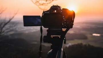 Kamera auf Stativ, filmend, vor Sonnenuntergang und Landschaft.