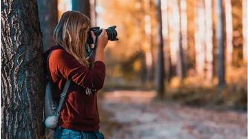 Frau fotografiert in herbstlichem Wald, Baum im Hintergrund.