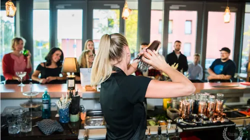 Bartender schüttelt Cocktails hinter der Bar; Gäste beobachten aufmerksam.