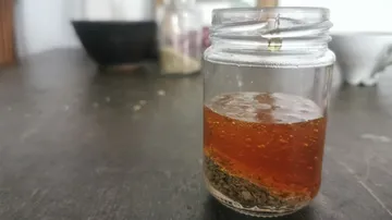 A glass of brown extract and herbs is on a table.