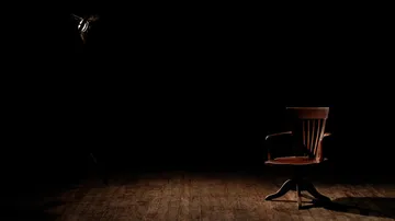 A wooden chair is placed under a spotlight in a darkened room.
