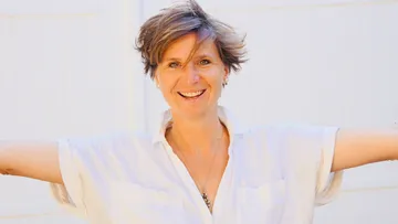 Eine Frau lächelt mit ausgebreiteten Armen vor einer Wand.