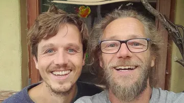 Two men smile and pose in front of a window.
