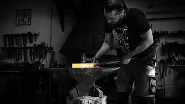 Blacksmith works glowing metal on anvil in workshop.