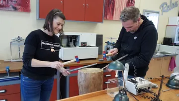 Zwei Personen bearbeiten einen Holzblock mit Werkzeugen.