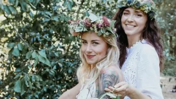 Two women with flower wreaths are smiling in nature.