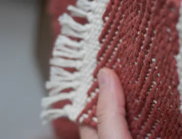 Hand holds woven, striped textile in reddish tones.