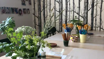 Table covered with plants and painting utensils in front of tree wallpaper.