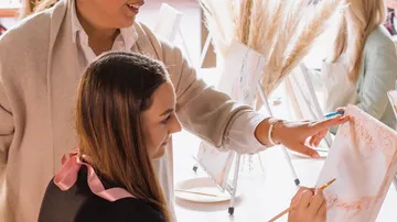 A woman paints while another gives instructions in a creative space.