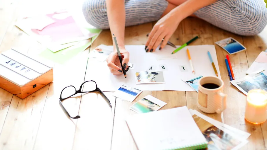 A person writes on paper surrounded by photos and pens.