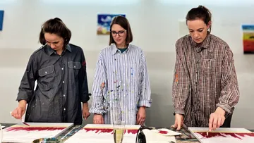 Three women paint canvases together in a studio.