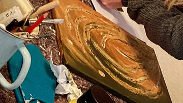 The artist paints on canvas, surrounded by painting supplies.