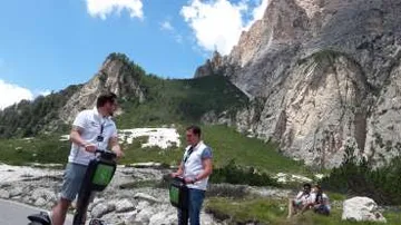 Zwei Männer stehen auf E-Scootern vor majestätischen Bergen.