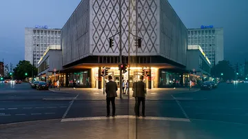 Mann steht an Ampel, reflektiert in einem Gebäude, Abenddämmerung.