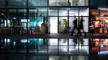 Zwei Personen gehen reflektiert an einem Glasgebäude vorbei.
