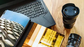 Books and camera lenses lie on a table next to a laptop.