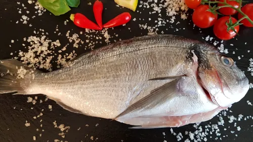 A fish is lying on a black stone surrounded by ingredients.
