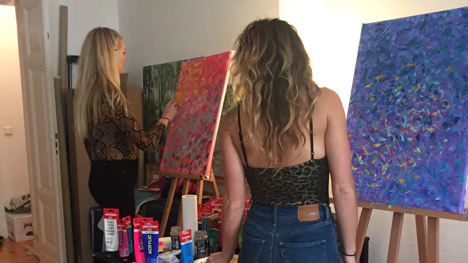 Two women are painting on canvases in a bright room.