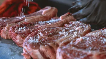Raw meat is sprinkled with salt, kitchen environment visible.
