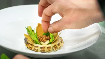Hand fügt Zutaten in ein kunstvolles, angerichtetes Gericht ein.
