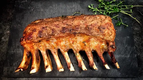 Lamb back roast lies on a dark surface with herbs.