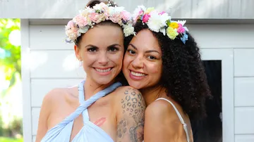 Two women are smiling, wearing flower wreaths, outdoors.