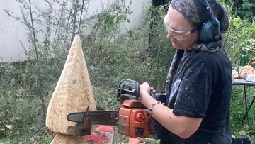 Wood artist carves an outdoor wooden sculpture with a chainsaw.