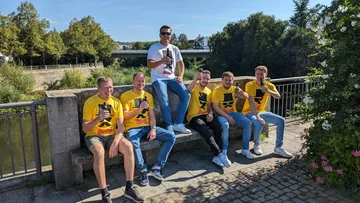 Sechs Männer in T-Shirts sitzen lächelnd an einem Fluss.