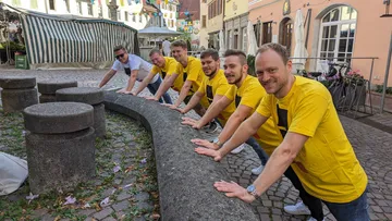 Six men in yellow T-shirts lean on a wall.
