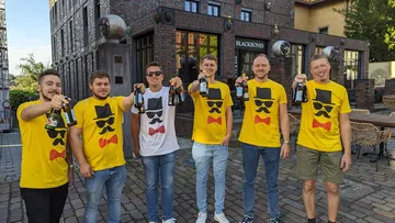 Sechs Männer in T-Shirts halten Bierflaschen vor einem Restaurant.