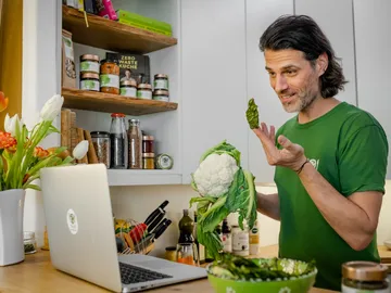 Mann hält Gemüse und spricht vor einem Laptop in der Küche.