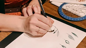 A hand draws plant patterns on paper at the table.