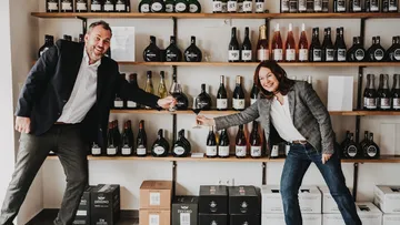 Two people toast with wine glasses in a wine warehouse.