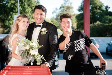 Bride and groom with flower bouquet pose happily in front of cars.
