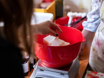 Eine Hand legt Seifenstücke in eine rote Schüssel auf einer Waage.