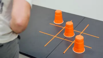Drei orangefarbene Becher stehen auf einem Tisch, Tic-Tac-Toe-Spiel.